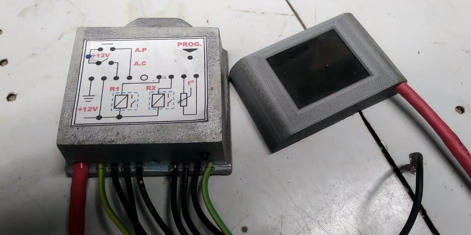 Sobre un banco de trabajo, una caja de aluminio fundido con una etiqueta de conexiones impresa en la tapa y siete cables saliendo por abajo, y al lado la carcasa gris impresa en 3D de la pantalla, con dos cables rojos gruesos