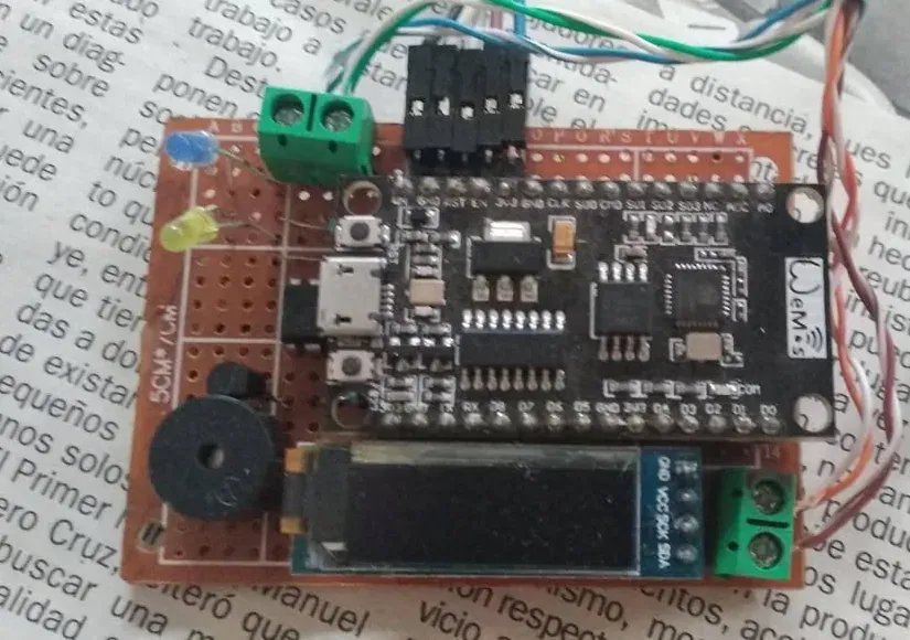 Placa perforada con una NodeMCU v3 soldada en el centro, una pantalla OLED alargada abajo, un zumbador negro, dos LED, dos bornes verdes y tiras de pines, con cables de colores saliendo por el borde