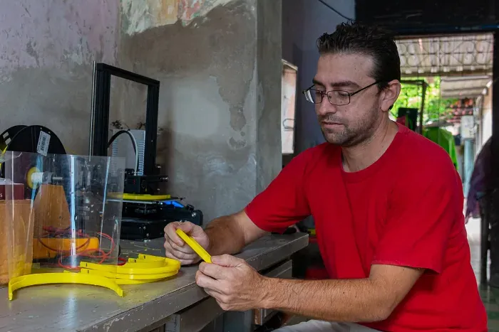 Un hombre sentado a un banco de trabajo repasa con los dedos una diadema de visera impresa en PLA amarillo; al lado hay una visera ya montada con su lámina de acetato y, al fondo, la impresora 3D con su rollo de filamento