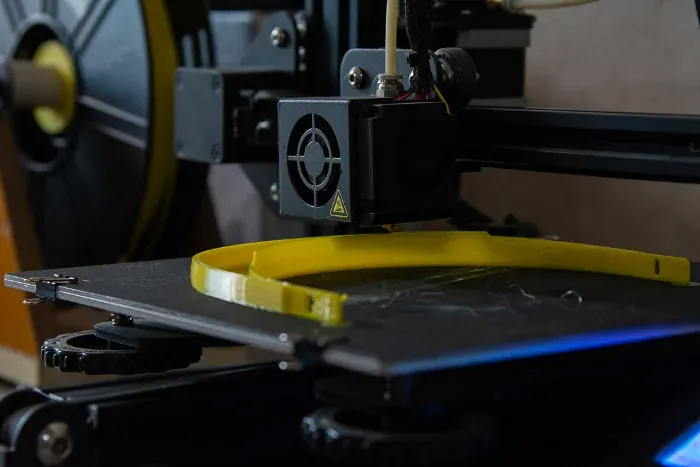 Primer plano del extrusor de una impresora 3D depositando filamento amarillo sobre la cama: la diadema de la visera está a medio imprimir, apoyada de canto